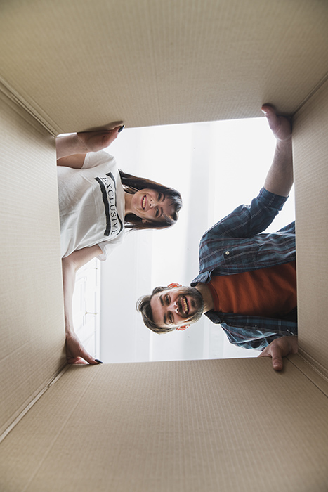 Una pareja sonríe al mirar dentro de una caja de cartón grande. La foto está tomada desde el interior de la caja, mostrando a la mujer a la izquierda y al hombre a la derecha, ambos asomándose desde arriba con alegría.