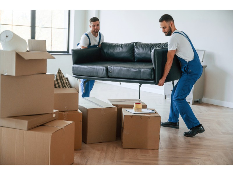Dos mudanceros trasladando un sofá en una habitación vacía rodeada de cajas.