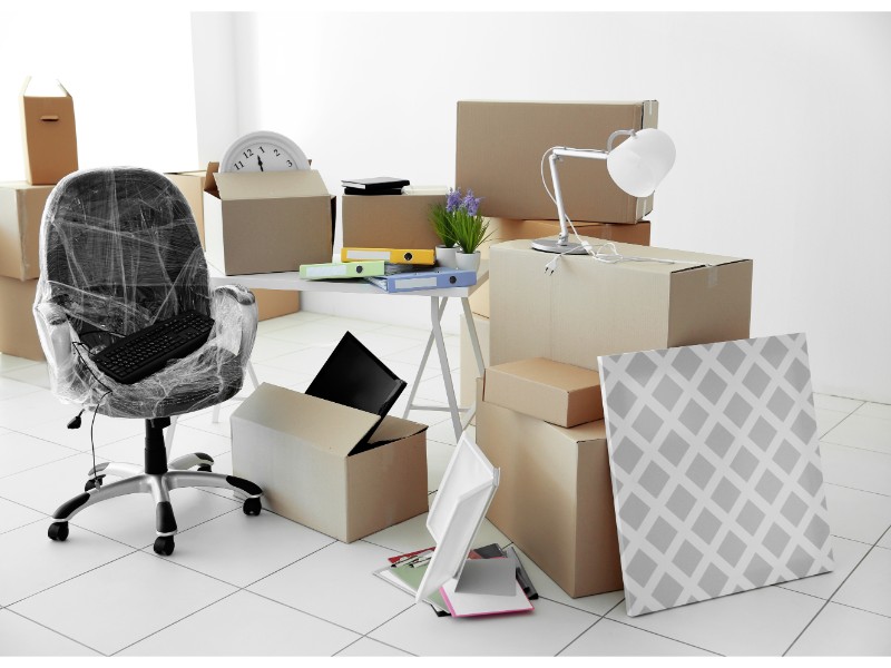 Un espacio de oficina con cajas de mudanza, una silla envuelta en plástico y objetos de trabajo, indicando una reubicación o mudanza