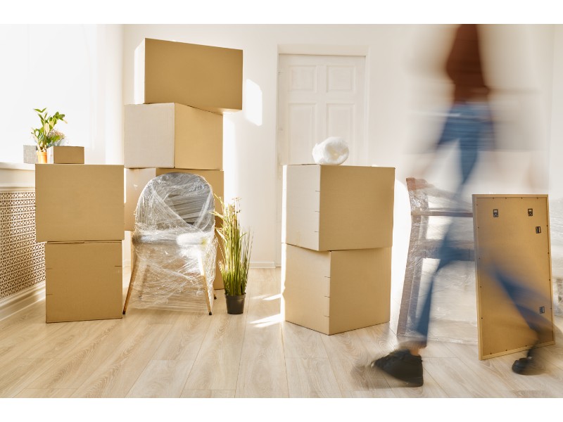 La imagen muestra una escena de mudanza dentro de un hogar. Se observan varias cajas de cartón apiladas y una silla cubierta con plástico, posiblemente esperando ser trasladada. Un objeto adicional también está en el suelo, y una persona aparece caminando de forma desenfocada en la parte derecha de la imagen, mientras transporta algún artículo.