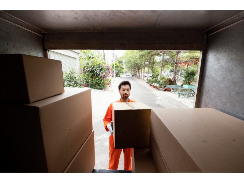 La imagen muestra a un trabajador con uniforme naranja y guantes, cargando una caja desde la parte trasera de un camión de mudanza, mientras que el entorno al aire libre se ve verde y tranquilo.