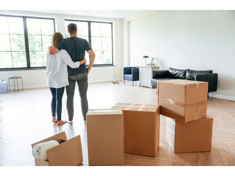 La imagen muestra a una pareja de pie en lo que parece ser su nuevo hogar, mirando hacia la habitación vacía. Están abrazados, con cajas de mudanza apiladas en primer plano. En el fondo, se puede ver que la habitación está parcialmente amueblada con un sofá negro y una mesa de escritorio. La pareja parece contenta y tranquila mientras observan el espacio.