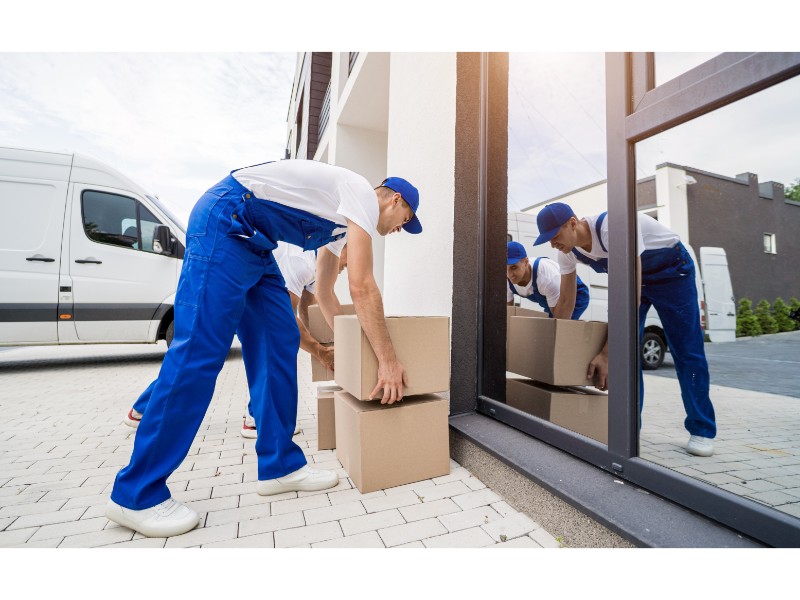 ChatGPT Plus La imagen muestra a un hombre vestido con un uniforme azul, levantando cajas de cartón afuera de un edificio, con una camioneta de mudanza aparcada detrás de él. También se refleja en la ventana del edificio
