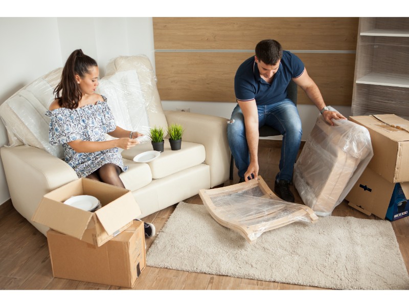 ChatGPT Plus La imagen muestra a dos personas, una mujer sentada en un sofá con una caja abierta que contiene platos, y un hombre frente a ella manipulando una silla envuelta en plástico, mientras están rodeados por más cajas y objetos.