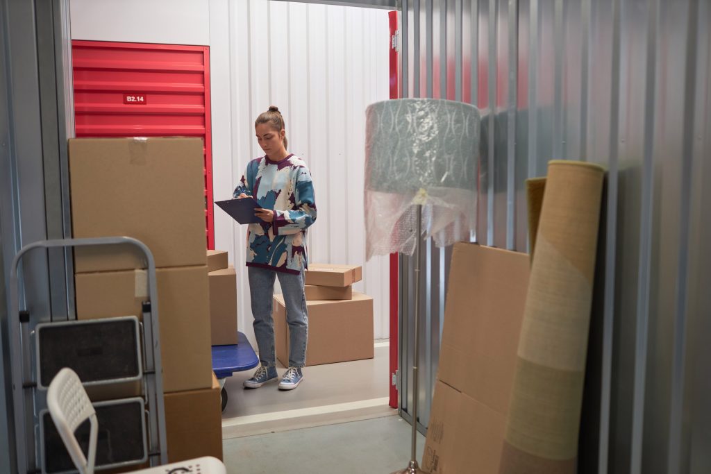 Mujer de pie en un almacén, frente a una unidad de almacenamiento con puerta roja, mientras sostiene un clip y toma notas. Hay varias cajas de cartón apiladas a su alrededor, y al fondo se ven alfombras enrolladas y un foco envuelto en plástico burbuja