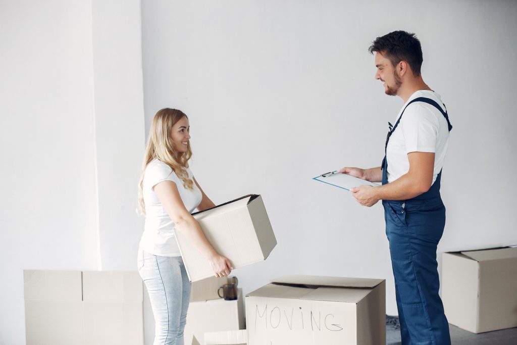 Una mujer sonriente sostiene una caja de mudanza mientras un hombre, vestido con overol, sostiene una carpeta con un formulario o lista. Ambos están en una habitación vacía rodeada de cajas etiquetadas con la palabra 'MOVING