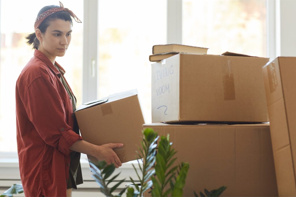 Mujer de pie junto a varias cajas de mudanza, sosteniendo una caja en las manos. Ella lleva una camisa roja y un pañuelo en el cabello, y al fondo se ven otras cajas etiquetadas como 'LIVING ROOM'. Hay algunas plantas en el primer plano