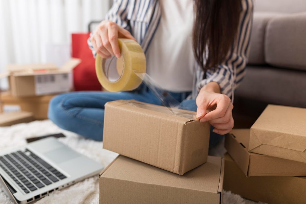 Mujer sentada en el suelo, tapando una caja de mudanza con cinta adhesiva. A su alrededor hay más cajas apiladas y una computadora portátil sobre una mesa. La mujer lleva una camisa a rayas y jeans.